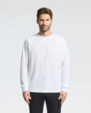 A person with short brown hair and a beard stands against a plain background, wearing the Psycho Bunny Men's Taylor Pique Long Sleeve Tee in white, paired with black pants, and looks directly at the camera with a neutral expression.