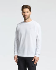A man with short brown hair and a beard stands against a plain backdrop wearing black pants and the Psycho Bunny "Mens Taylor Pique Long Sleeve Tee" (B6T580F200), crafted from white piqué cotton with an embroidered bunny logo on the left chest.