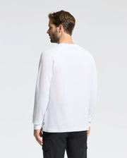 A person with short hair, facing away from the camera, wears a white Psycho Bunny MENS TAYLOR PIQUE LONG SLEEVE TEE made of soft Pima cotton and black pants against a plain neutral background.