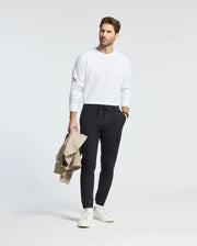 A man in a Psycho Bunny white Taylor Pique long sleeve tee and black jogger pants holds a beige jacket. He wears white sneakers, standing stylishly against a plain background with understated charm.