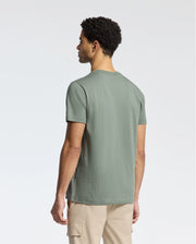 A person with short curly hair stands with their back to the camera, wearing a Psycho Bunny MENS CLASSIC CREW NECK TEE in green Pima cotton and beige pants against a plain white background.