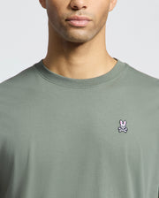 A person models Psycho Bunny's MENS CLASSIC CREW NECK TEE (B6U014B200) in light green Pima cotton, with a pink bunny-and-crossbones logo on the chest. The image is cropped to highlight the upper torso and part of the face against a plain backdrop.