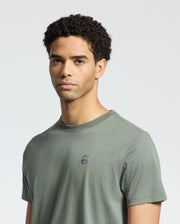 A person with short, curly hair is wearing the Psycho Bunny MENS CLASSIC CREW NECK TEE - B6U014B200 in gray-green Pima cotton, featuring an embroidered skull and crossbones on the chest. The regular fit and light background make them stand out.