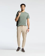 A person poses with a jacket over one shoulder, wearing the Psycho Bunny MENS CLASSIC CREW NECK TEE in green Pima cotton, paired with beige cargo pants and brown shoes. The plain white background highlights their relaxed stance and regular fit style.
