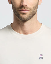 A man is wearing a Psycho Bunny MENS CLASSIC CREW NECK TEE - B6U014B200 in light beige, made from premium jersey. It has a small embroidered logo featuring a pink-eared bunny with a skull below it on the left chest area. The background is plain light gray, showing only his lower face and chest.