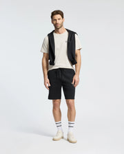 A man stands against a plain background wearing a Psycho Bunny MENS CLASSIC CREW NECK TEE (B6U014B200) in white, paired with black shorts and a black sweatshirt draped over his shoulders. His look is completed with striped socks and white sneakers.