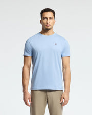 A person wearing a Psycho Bunny MENS CLASSIC CREW NECK TEE, style B6U014B200, in light blue Pima cotton with an embroidered chest logo and beige pants stands against a plain white background.