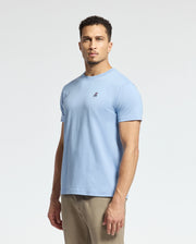 A man is wearing a Psycho Bunny MENS CLASSIC CREW NECK TEE B6U014B200 in light blue, featuring a small chest logo, paired with beige pants, against a plain white background.