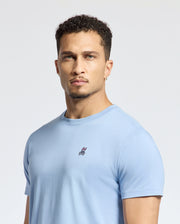 A person with short curly hair and a goatee wears a Psycho Bunny MENS CLASSIC CREW NECK TEE - B6U014B200 in light blue, crafted from soft Pima cotton, featuring a small embroidered chest logo. The background is plain white.
