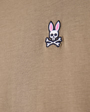 Close-up of a khaki tee made from 100% Pima cotton, showcasing the Psycho Bunny embroidered logo with a cartoonish skull and crossbones design featuring rabbit ears.