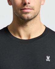 Someone is wearing a Psycho Bunny MENS CLASSIC CREW NECK TEE - B6U014CRPC in black, made from 100% Pima cotton. It has a small embroidered rabbit logo with crossed bones on the chest. The plain, light-colored background accentuates the tee's design and taped seams.