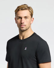 A person with short blonde hair wears a Psycho Bunny MENS CLASSIC CREW NECK TEE - B6U014CRPC, made of 100% Pima cotton, with taped seams and a small chest logo. They're set against a plain white background.