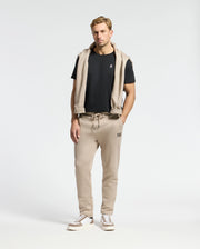 A man in Psycho Bunny's black MENS CLASSIC CREW NECK TEE and beige joggers stands against a plain background. A matching beige hoodie with taped seams drapes over his shoulders. He sports white sneakers with brown accents, and his right hand rests in his pocket.