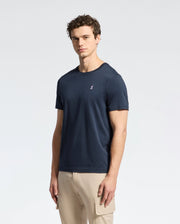 A man with short curly hair wears a Psycho Bunny MENS CLASSIC CREW NECK TEE - B6U014CRPC in navy blue, made from 100% Pima cotton, featuring a small chest logo, paired with beige cargo pants and standing against a light background.