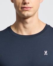 A person wears the Psycho Bunny MENS CLASSIC CREW NECK TEE (B6U014CRPC) in dark 100% Pima cotton, featuring a small pink bunny with crossbones on the left chest. Only their lower face, neck, and upper torso are visible.