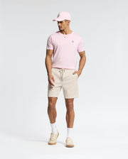 A man wears a light pink Psycho Bunny pima cotton cap and matching MENS CLASSIC CREW NECK TEE - B6U014CRPC, paired with beige drawstring shorts, white socks, and beige sneakers. Against a plain background, he looks down with his hands at his sides.