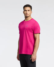 A man in a bright pink Psycho Bunny MENS CLASSIC CREW NECK TEE - B6U014CRPC with an embroidered bunny logo on the chest and black pants stands against a light background. His short curly hair frames his serious expression, giving him a thoughtful look.