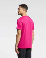 A man in a vibrant pink Psycho Bunny MENS CLASSIC CREW NECK TEE - B6U014CRPC and black pants stands sideways against a light gray backdrop, highlighting the embroidered bunny logo's subtle elegance.