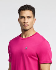 A person with short curly hair, in a Psycho Bunny MENS CLASSIC CREW NECK TEE - B6U014CRPC, vibrant pink and with an embroidered bunny logo, stands against a plain white background. They wear gold hoop earrings and have a neutral expression.