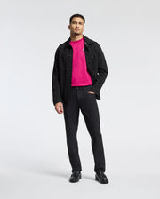 A man with short hair and a beard, standing against a plain background, wears a bright pink Psycho Bunny Classic Crew Neck Tee (B6U014CRPC) under a black jacket with matching pants and shoes. One hand in his pocket complements the ensemble, highlighted by subtle embroidered Bunny logo details.