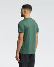 A man with short curly hair, wearing a green Psycho Bunny MENS CLASSIC CREW NECK TEE (B6U014F200) and black pants, stands with his back to the camera against a plain light background.
