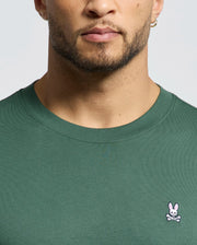 A man wears the Psycho Bunny MENS CLASSIC CREW NECK TEE (B6U014F200) in green, featuring a small pink bunny and white crossbones logo on the left chest. Only his lower face, neck, and upper chest are shown.