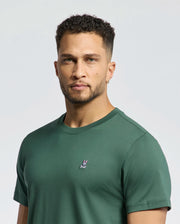 A man with short curly hair and a trimmed beard wears the Psycho Bunny MENS CLASSIC CREW NECK TEE (B6U014F200) in green, featuring a small bunny logo on the chest, standing against a plain light background.