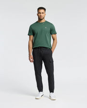 A man with short hair and a trimmed beard stands against a white background, wearing Psycho Bunny's MENS CLASSIC CREW NECK TEE - B6U014F200 in dark green, paired with black cargo pants and white sneakers, hands relaxed by his sides.