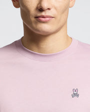 A person wearing the Men's Classic Crew Neck Tee in light pink from Psycho Bunny demonstrates its versatility, featuring a small embroidered bunny with crossed bones logo on the chest. This 100% pima cotton tee highlights their upper torso against a neutral background.