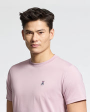 A person in a versatile light pink Psycho Bunny MENS CLASSIC CREW NECK TEE - B6U014F200, made from 100% pima cotton and featuring a small embroidered rabbit on the chest, stands against a plain white background with short styled dark hair and a neutral expression facing the camera.