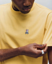 A person in a light yellow Psycho Bunny MENS CLASSIC CREW NECK TEE (B6U014F200) with a small pink-eared bunny logo above crossed bones on the chest gently pulls the sleeve, face partially visible.