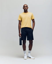 A man stands against a plain background wearing the Psycho Bunny MENS CLASSIC CREW NECK TEE - B6U014F200 in yellow, black shorts, white socks with black stripes, white sneakers, holding a black water bottle in his right hand.