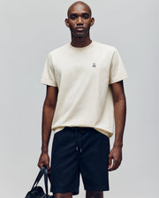 A man stands against a plain background wearing the Psycho Bunny MENS CLASSIC CREW NECK TEE - B6U014F200, a light beige, regular-fit pima cotton tee with an embroidered bunny logo on the chest, paired with black drawstring shorts and holding a black bag.