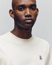 A man with a shaved head wears a cream Psycho Bunny MENS CLASSIC CREW NECK TEE (B6U014F200), featuring regular fit pima cotton and a small pink bunny and crossbones logo on the chest, looking at the camera with a neutral expression.