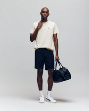 A man stands against a plain background wearing the Psycho Bunny MENS CLASSIC CREW NECK TEE - B6U014F200 in cream, paired with black shorts, white socks, white sneakers, holding a black duffel bag and touching his neck.
