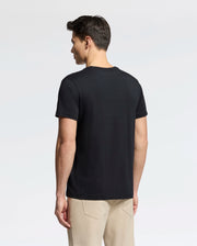A man faces away from the camera, dressed in a plain black Psycho Bunny Men's Classic V Neck Tee (B6U100ARPC) and beige pants against a simple white background.