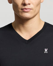 A person is wearing a Psycho Bunny Men's Classic V-Neck Tee (B6U100ARPC) in black, showcasing the pink bunny head and crossbones logo on the left side. The image is cropped to display only the upper torso and part of the face.