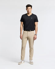 A man exudes confidence in a sleek black Psycho Bunny MENS CLASSIC V NECK TEE - B6U100ARPC, crafted from soft Pima cotton. Paired with beige pants and white sneakers, he embodies effortless style, his wrist adorned with a watch as he gazes forward against a plain background.