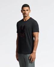 A man confidently poses against a plain background, wearing the Psycho Bunny MENS PIERCE EMBROIDERED GRAPHIC TEE - B6U115E200. The black tee, crafted from soft Pima cotton, showcases an eye-catching red embroidered design of a skull with bunny ears. With short hair and gray pants, he gazes directly at the camera.