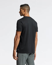 A man with short hair stands against a plain white background, facing away from the camera. He is wearing the Psycho Bunny MENS PIERCE EMBROIDERED GRAPHIC TEE - B6U115E200 made of soft Pima cotton and gray pants.