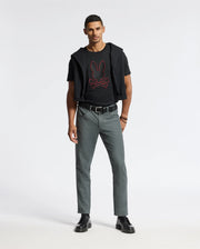 A man stands confidently against a plain background wearing the Psycho Bunny Men's Pierce Embroidered Graphic Tee - B6U115E200 in black, showcasing red bunny embroidery. He complements it with gray pants and black shoes, with a black Pima cotton sweater elegantly draped over his shoulders. His hands rest at his sides, completing the stylish ensemble.