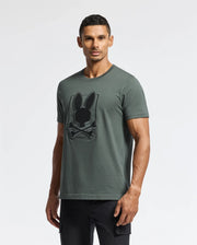 A man dressed in a green Psycho Bunny MENS PIERCE EMBROIDERED GRAPHIC TEE, featuring bunny embroidery and made from soft Pima cotton, stands against a plain white backdrop. He has short, dark hair and a beard, with his hands relaxed by his sides while wearing black pants.