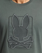 Detailed view of a dark green Psycho Bunny Men's Pierce Embroidered Graphic Tee (B6U115E200) crafted from soft Pima cotton, showcasing an embroidered design of a bunny head with crossed bones beneath. The embroidery features an outline, creating a raised texture on the fabric.
