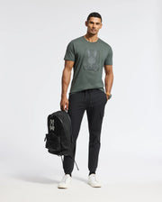 A man confidently stands in black joggers wearing a Psycho Bunny's Men's Pierce Embroidered Graphic Tee, crafted from Pima cotton and decorated with bunny embroidery. He holds a black backpack adorned with the brand's logo while sporting white sneakers, set against a plain white background.