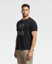 A man with short, curly hair wears the Psycho Bunny men's MONARC Front Bunny Graphic Tee, featuring a bunny logo above crossbones. Made from soft Pima cotton, it pairs perfectly with his beige pants against the light gray background.