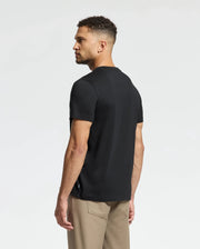 A man with short curly hair, wearing a black Psycho Bunny MENS MONARC FRONT BUNNY GRAPHIC TEE - B6U211E200 in soft Pima cotton and khaki pants, stands sideways glancing left against a plain light gray background.