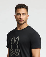A man with short curly hair and a beard wears the MENS MONARC FRONT BUNNY GRAPHIC TEE - B6U211E200 by Psycho Bunny, made of soft Pima cotton and featuring a large stylized bunny with crossed bones. He stands against a plain white background.