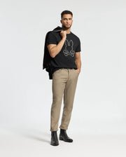 A man stands against a plain background, wearing a black Psycho Bunny Tee (B6U211E200), beige pants, and black boots, with a Monarc jacket slung over his shoulder, creating a relaxed yet stylish look.