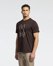 A man wears a Psycho Bunny MONARC Front Bunny Graphic Tee made from 100% Pima cotton, showcasing a rabbit-and-crossbones design. He poses against a white background, gazing at the camera in stylish khaki pants.