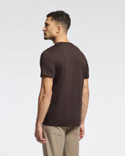 A man with short curly hair wearing a Psycho Bunny MENS MONARC FRONT BUNNY GRAPHIC TEE, crafted from 100% Pima cotton, and beige pants stands with his back to the camera against a plain white background.